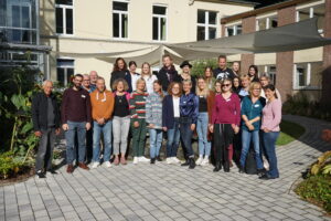 Ein Gruppenbild. Knapp 30 Personen unterschiedlichen Alters und Geschlechts stehen versammelt vor einem Seminargebäude und blicken in die Kamera. Die Sonne scheint.