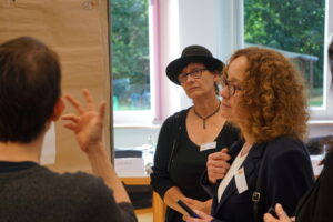 Eine Szene aus einem Workshop. Fokussiert wird hier eine Frau mit Brille, die gerade aufmerksam einer anderen Person zuzuhören scheint. Sie trägt eine Brille, einen schwarzen Hut und ein schwarzes Oberteil. Im Hintergrund ist durch ein großes Fenster unscharf ein Garten zu sehen.